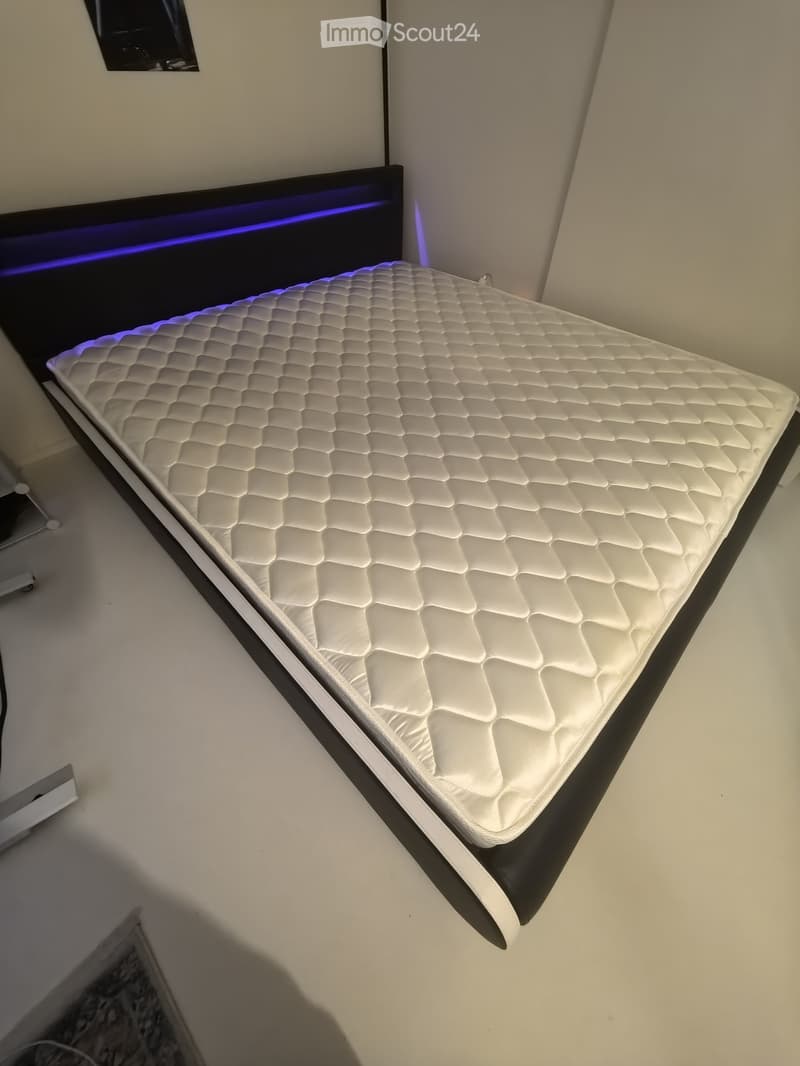 A modern bedroom with a white quilted mattress on a black bed frame, illuminated by blue LED lighting along the headboard.