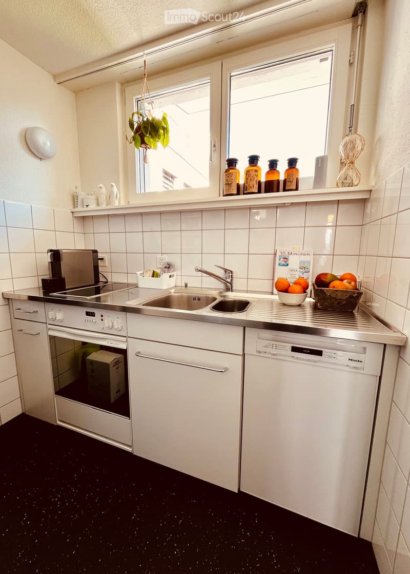 Modern kitchen with white cabinets, stainless steel appliances, and a black speckled floor. Features include a sink, oven, dishwasher, coffee maker, and various decorative items on the windowsill.