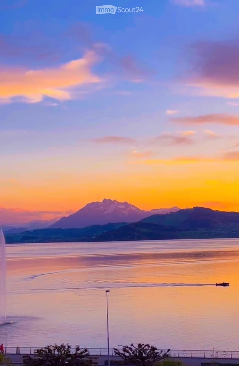 A scenic view of a lake at sunset with a fountain, mountains in the background, and a few people walking along the waterfront.