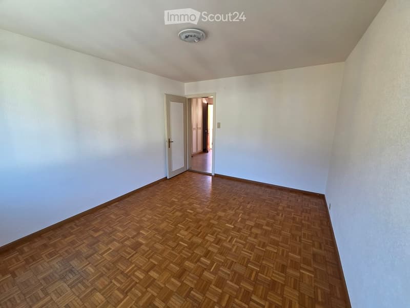Empty room with light-colored walls and wooden parquet flooring, featuring a single door leading to another room.