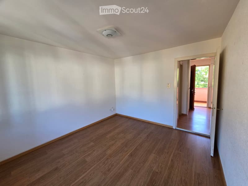 Empty room with light-colored walls and wooden flooring, featuring a ceiling light fixture and an open doorway leading to another area.
