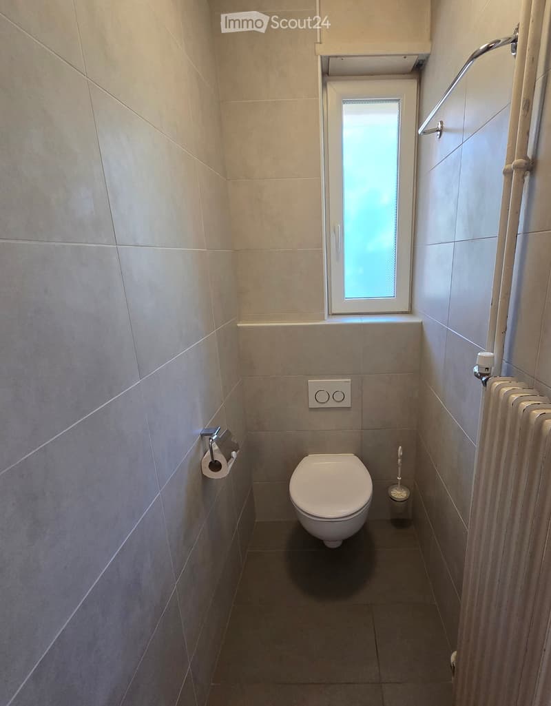 Small bathroom with a toilet, toilet paper holder, toothbrush holder, and a radiator. Light-colored tiled walls and a window above the toilet.