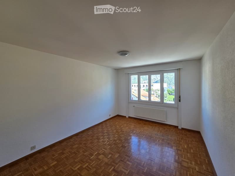 Empty room with wooden parquet flooring, white walls, and a large window with a radiator below it.