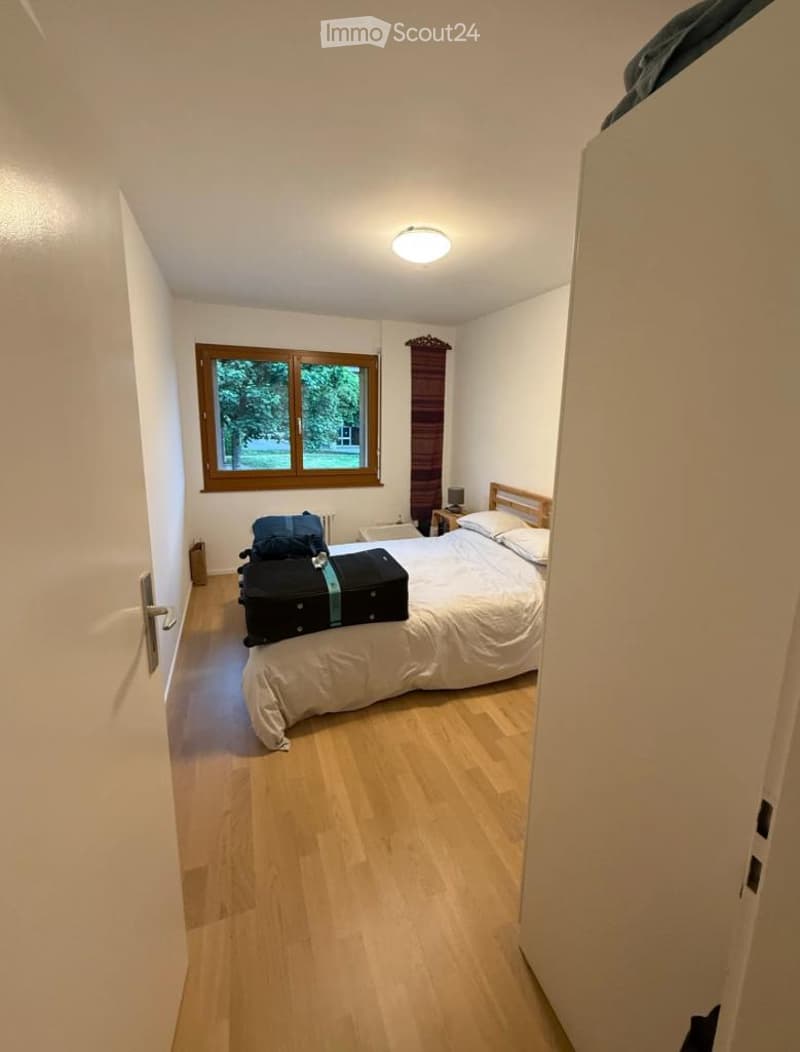 Cozy bedroom with light wooden flooring, a large window with wooden frame, a single bed with white bedding, a nightstand with a lamp, and luggage placed on the floor.