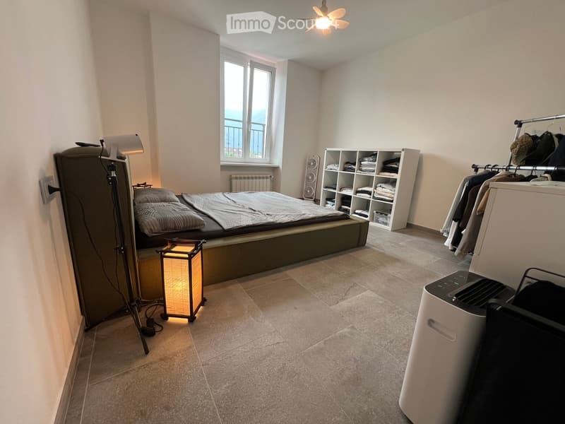 A modern bedroom with a large bed, bedside table, lamp, shelf with folded clothes, speaker, clothing rack, and air conditioner. The room has tiled flooring and large windows with a view.