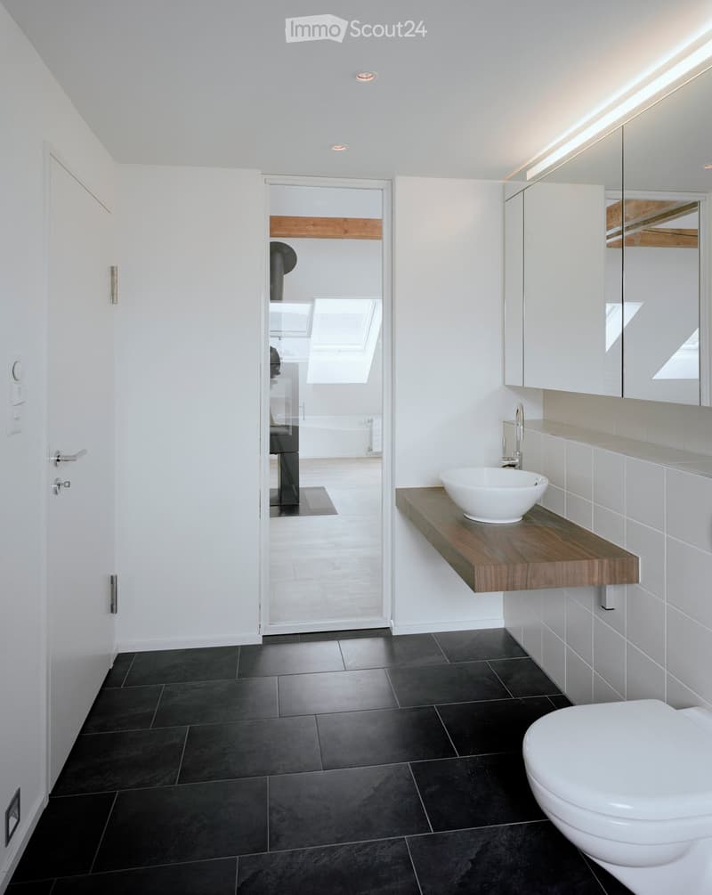 Modern bathroom with black tiled floor, white walls, a floating wooden sink, a toilet, and a large mirror with built-in lighting. A glass door leads to another room.