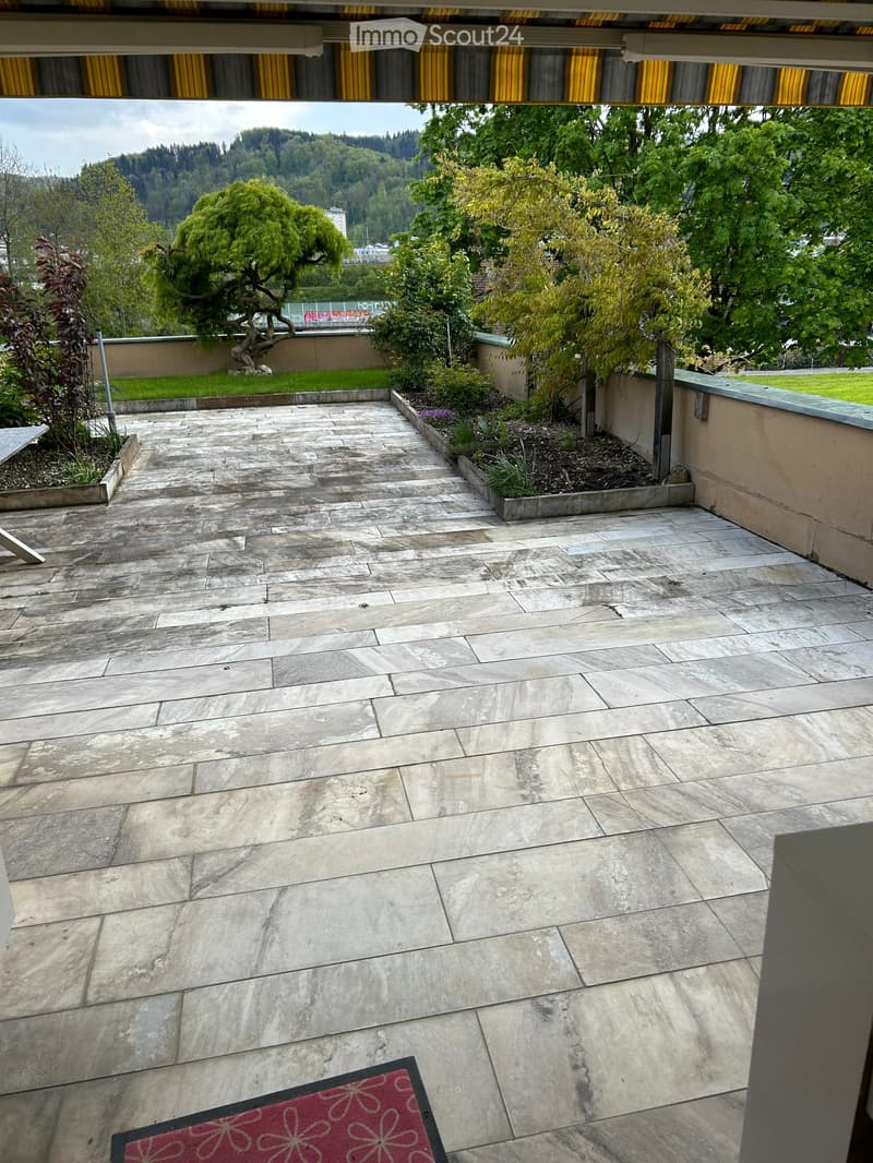 Outdoor patio with stone tiles, small garden beds, trees, and a scenic view of a hilly landscape.
