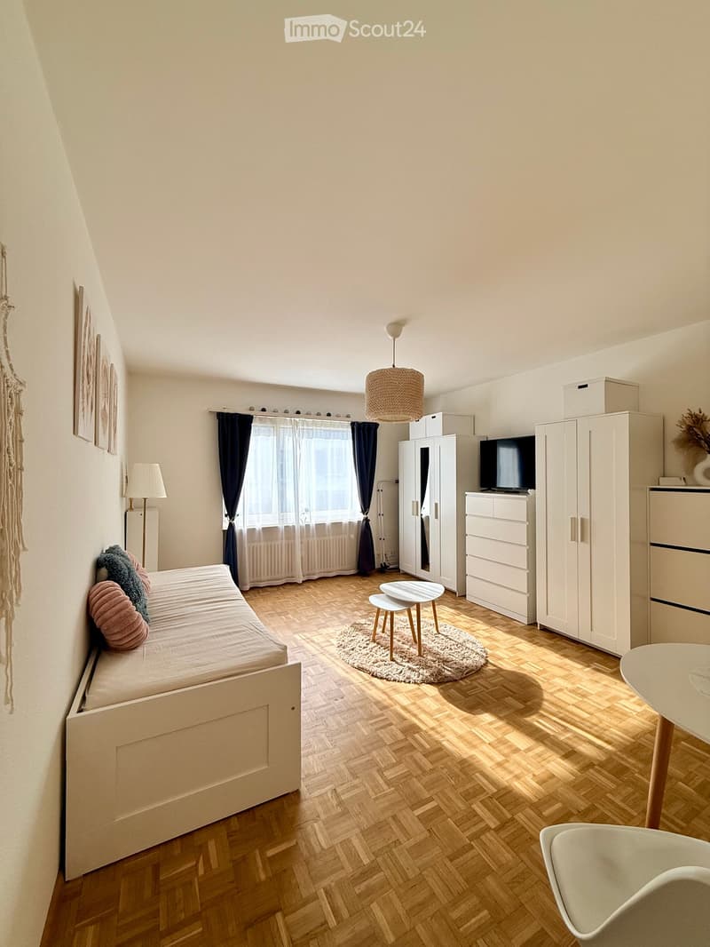 Cozy bedroom with a wooden bed, small table, chair, wardrobe, and a hanging lamp. The room features wooden flooring, a window with curtains, and decorative elements like a picture frame and a woven wall hanging.