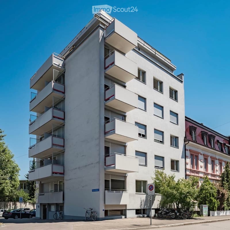 Modern multi-story apartment building with balconies, white facade, and multiple windows. The building is surrounded by trees and parked vehicles.