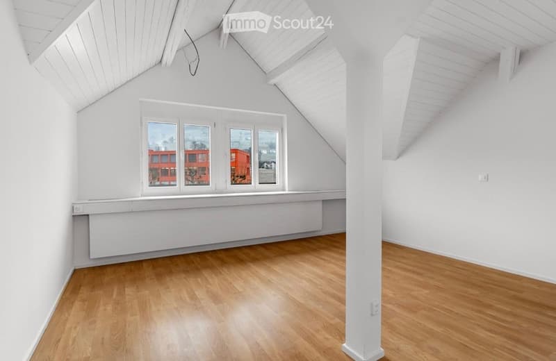 Empty attic room with wooden flooring, white walls, and large windows offering a view of a red building outside.