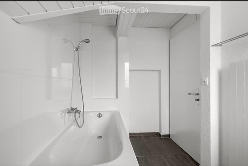 Modern white bathroom with a bathtub, showerhead, and faucet. The space features white tiled walls, a dark tiled floor, and a white door.