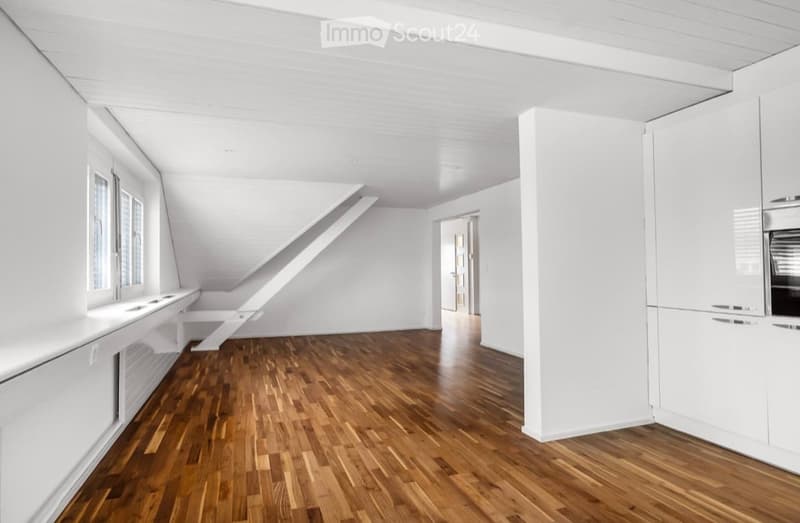 Bright, empty room with wooden flooring, white walls, and large windows. The space features a slanted ceiling and a doorway leading to another area.