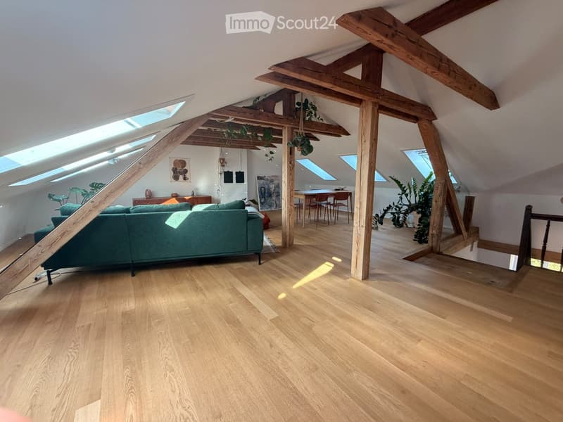 Open-plan attic space with wooden beams, skylights, hardwood flooring, green sofa, dining table, chairs, and artwork.