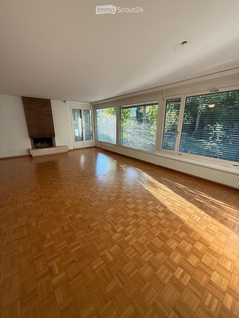Empty room with wooden herringbone flooring, large windows with blinds, and a fireplace against one wall.