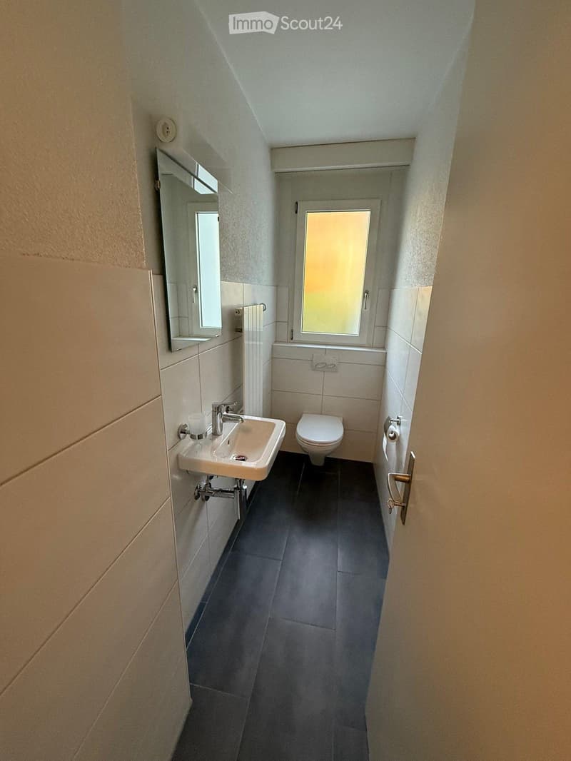 Compact bathroom with a white sink, chrome faucet, toilet, and a small mirror above the sink. The room has light-colored tiles and a dark floor.