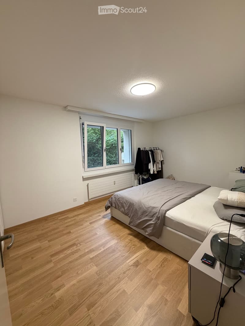 Cozy bedroom with wooden flooring, a double bed with grey bedding, a nightstand with a lamp, and a clothing rack with hanging garments.