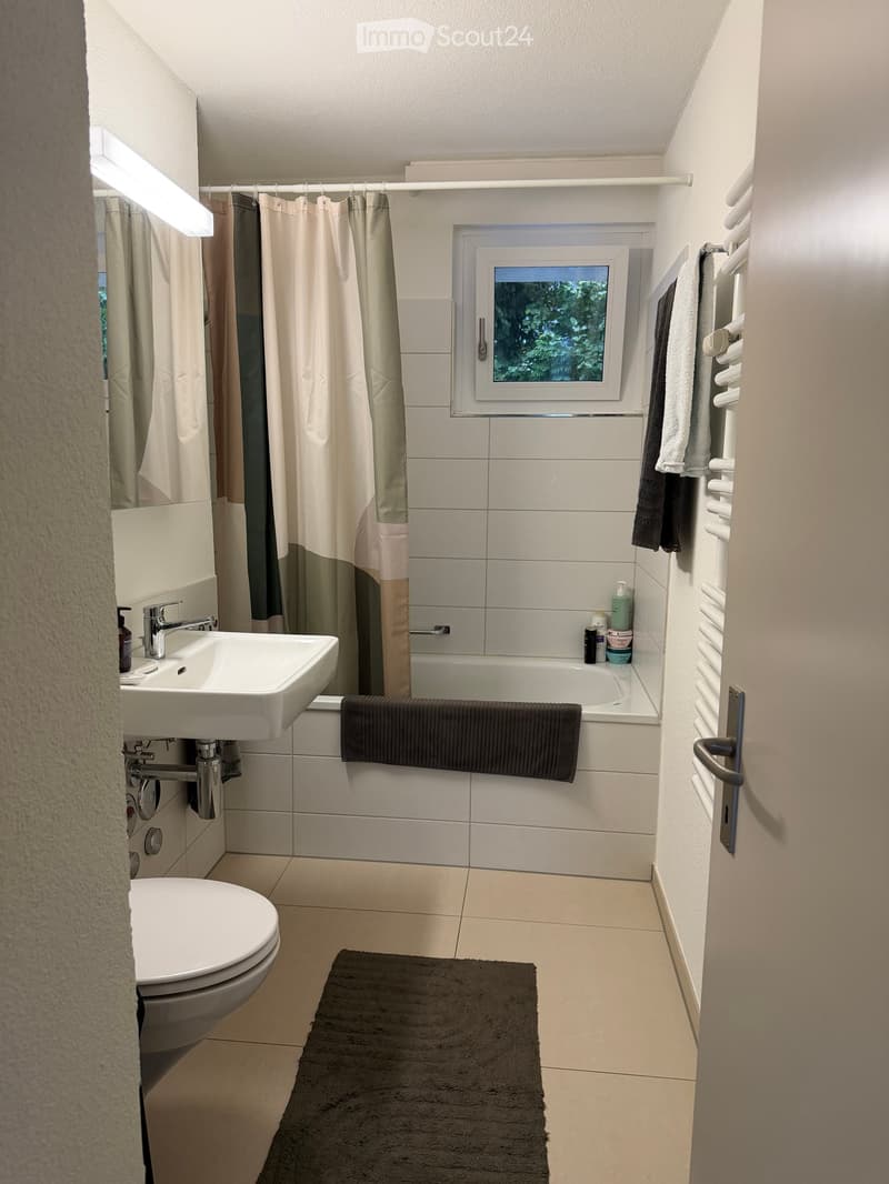 Modern bathroom with white tiles, a bathtub, toilet, sink, and a window with a view of greenery. Includes towels, a rug, and a radiator.