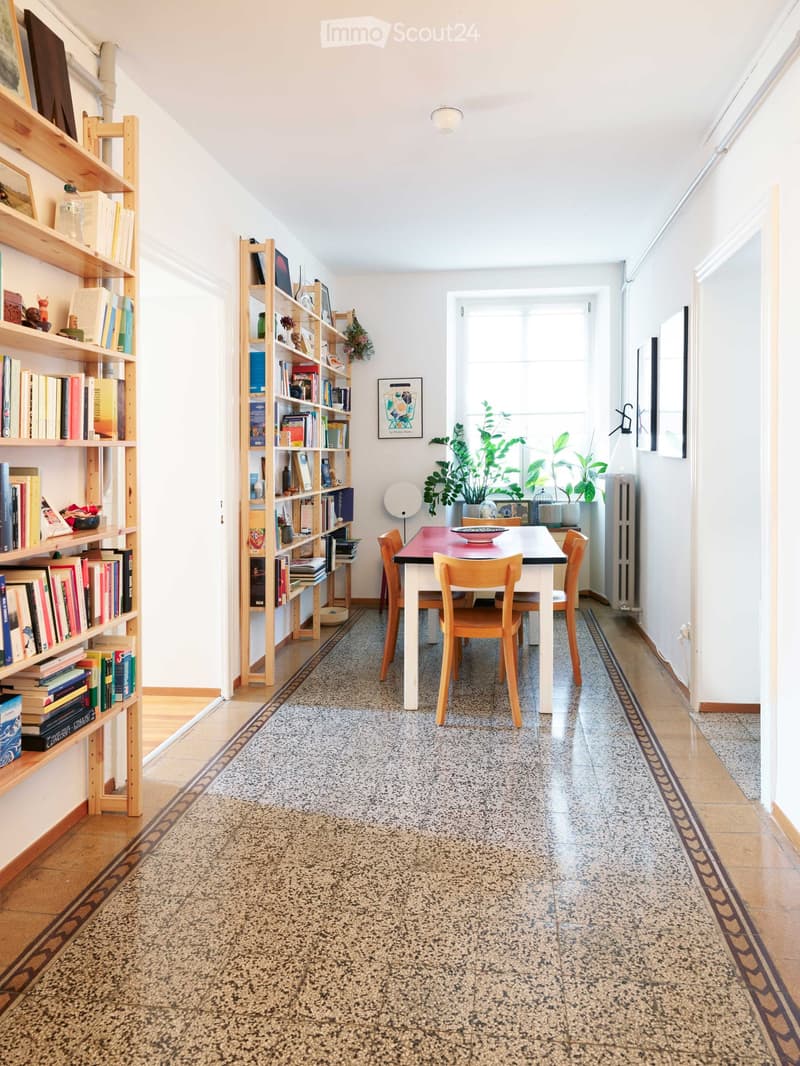 A cozy, well-lit room with a dining table and chairs, wooden bookshelves filled with books and decorative items, and a large window with plants on the sill.