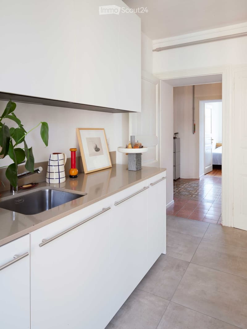 Modern kitchen with white cabinets, stainless steel sink, and a countertop with decorative items including a framed picture, vase, and bowl. A doorway leads to another room with visible furniture.