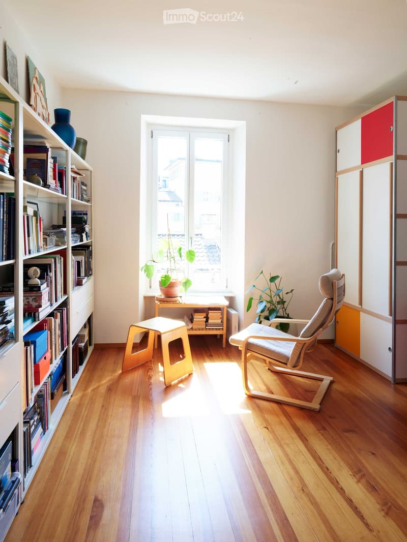 Bright, minimalist room with wooden flooring, a large window, a bookshelf filled with books and decorative items, a small table with a plant, a modern chair, and a small stool.