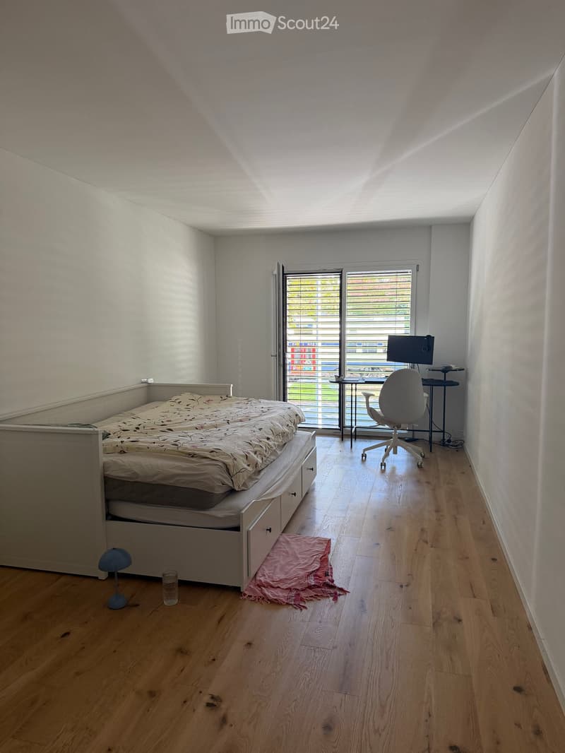 A modern bedroom with a bed, a desk setup including a chair, table, and screen, a small lamp, and a cup on the floor. The room features wooden flooring and large windows with blinds.