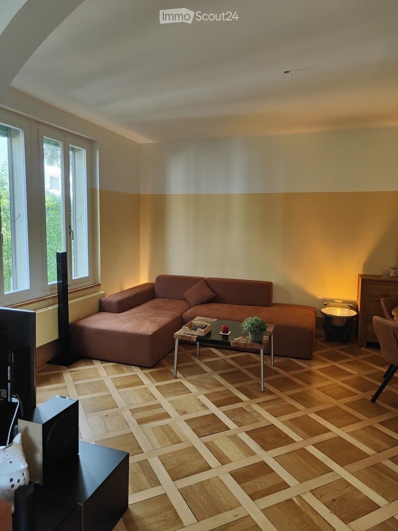 Living room with wooden flooring, large window, brown sectional sofa, coffee table with a small plant, and a chair.