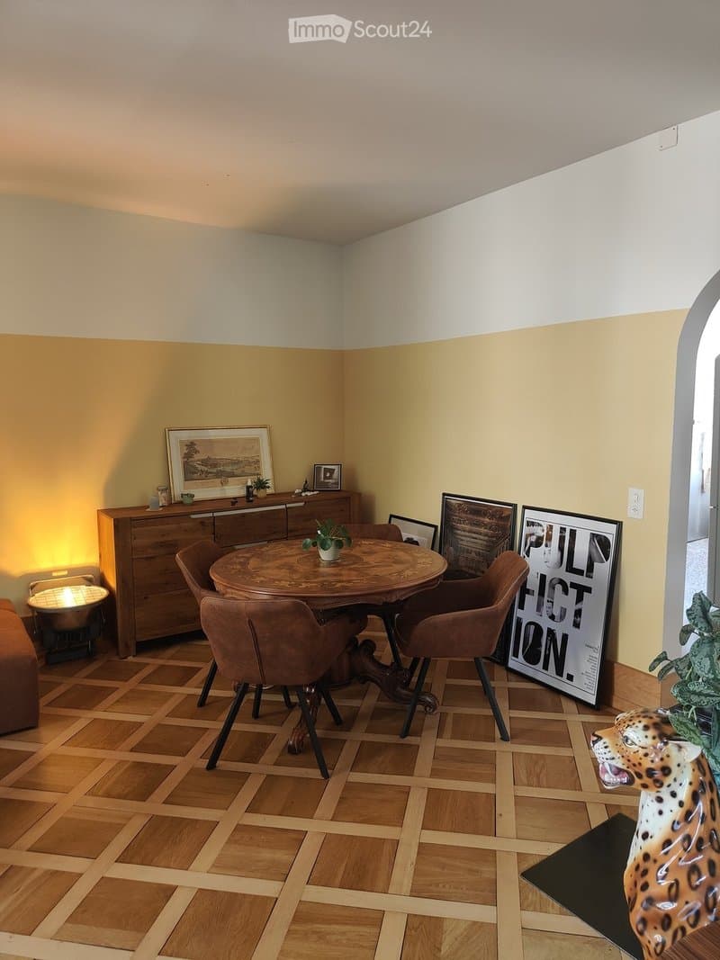 A cozy dining area with a round wooden table surrounded by four brown chairs, a wooden sideboard with a picture frame and lamp, and a large poster on the wall. The floor is made of wooden tiles, and there is a leopard statue on the right side.