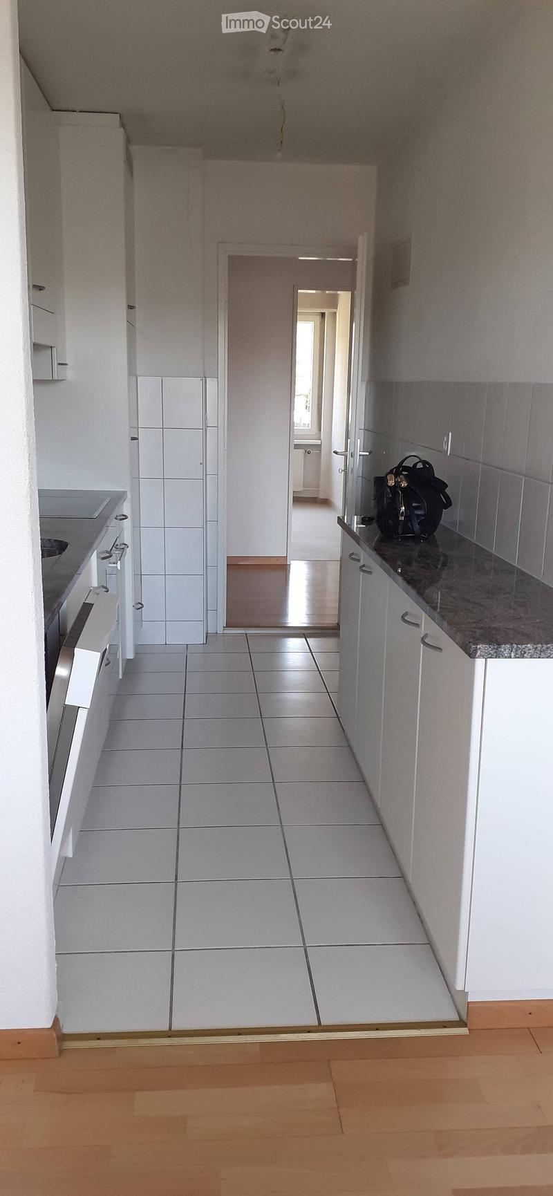 A clean, modern hallway with white tiled walls, wooden flooring, and a view into a kitchen area with white cabinets and a granite countertop.