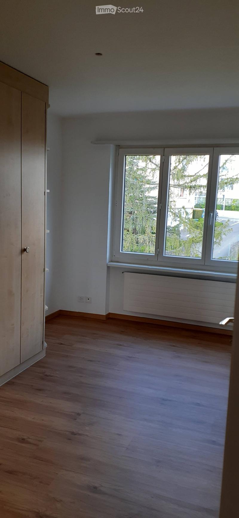Empty room with wooden flooring, white walls, a window with a radiator below, and a door on the left side.