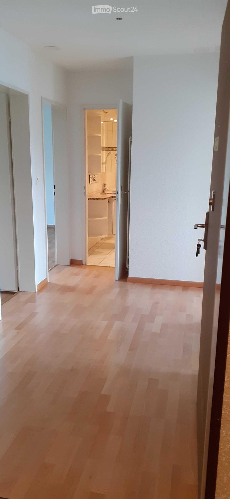 Empty hallway with light wooden flooring, white walls, and open doors leading to a bathroom and another room.