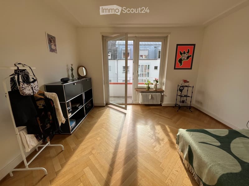 Bright bedroom with wooden herringbone flooring, large window with open door leading to balcony, bed with green and white patterned bedspread, wardrobe, mirror, small table with vase and picture frame, radiator, and clothing rack.
