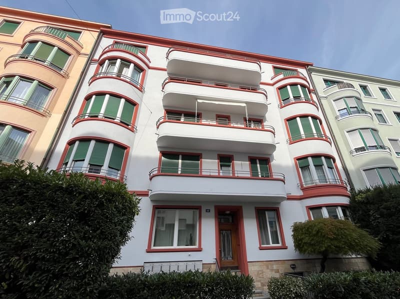 Multi-story residential building with white facade, red trim, and multiple balconies with green shutters.