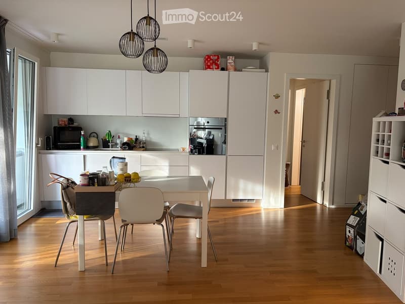 Modern kitchen with white cabinets, a dining table with chairs, and various appliances including a microwave, oven, and refrigerator. The space features wooden flooring and pendant lighting.