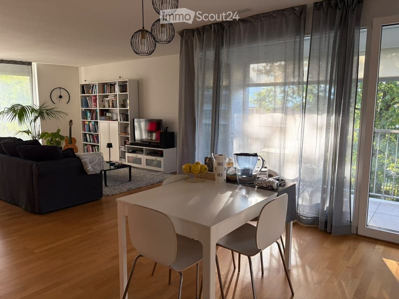 Bright living and dining area with wooden flooring, large windows, and a balcony access. Features a sofa, dining table with chairs, bookshelf, television, and decorative items.