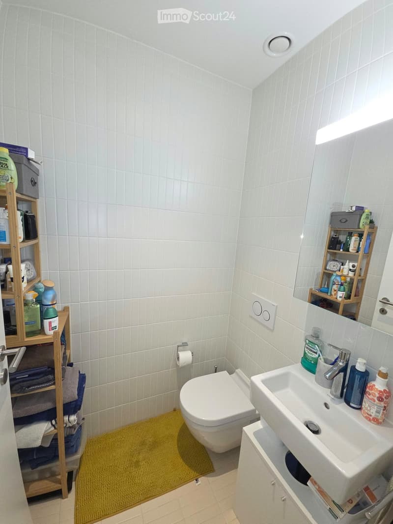 A compact bathroom with white tiled walls and floor, featuring a toilet, sink with a mirror, shelves with toiletries, and a yellow bath mat.