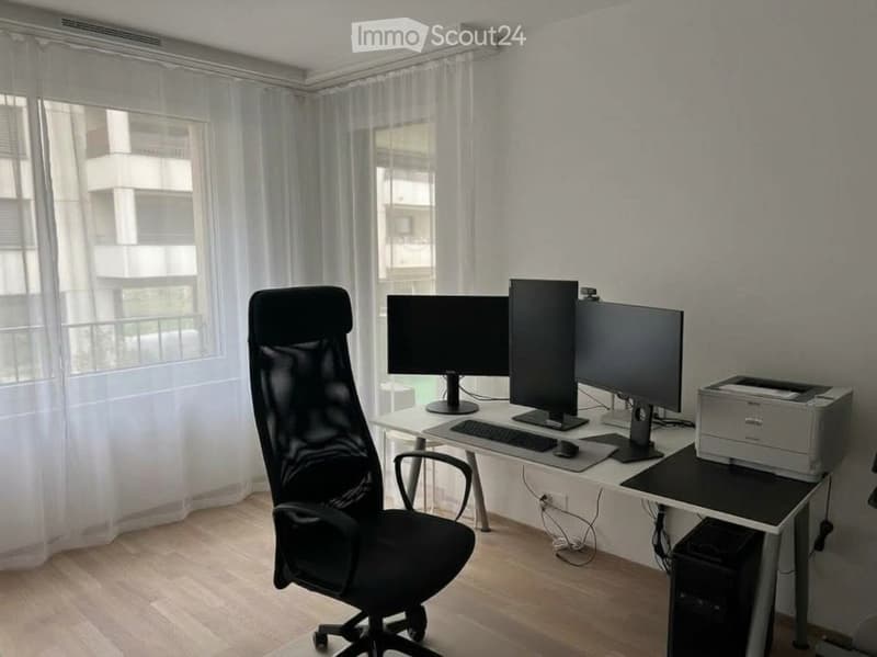 A modern office space with a large desk, multiple computer monitors, a comfortable office chair, and a printer. The room features large windows with sheer curtains and wooden flooring.