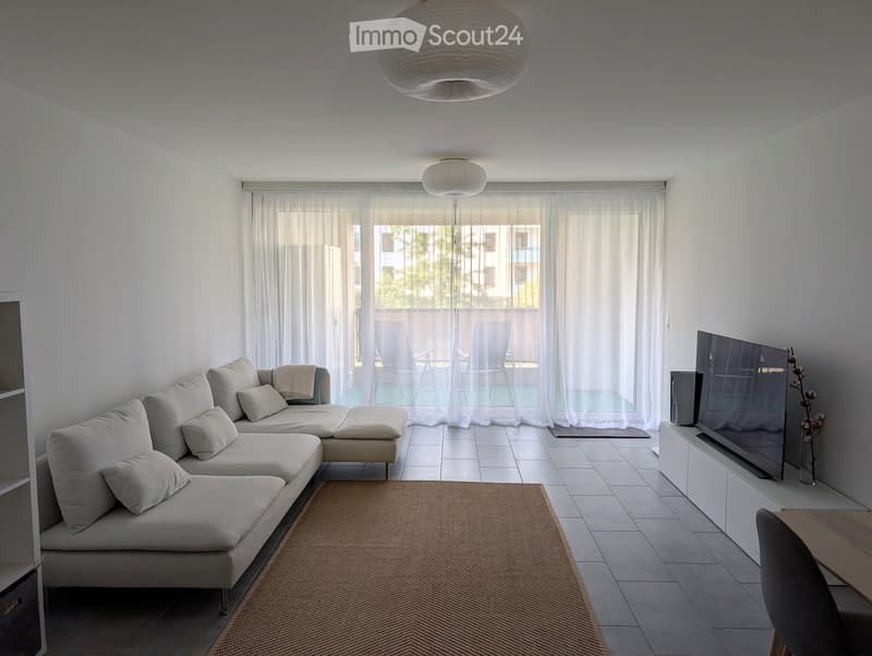 Modern living room with a white sofa, coffee table, TV, and a view of a balcony area with outdoor furniture.