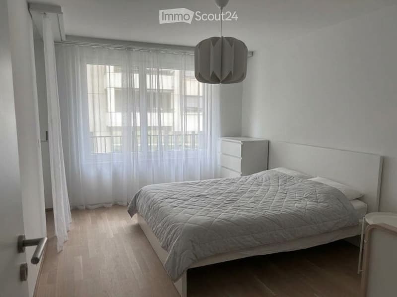 Minimalist bedroom with a white bed, grey bedding, a nightstand, and a modern hanging lamp. The room features light wooden flooring and large windows with sheer curtains.