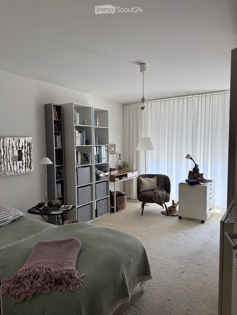 Cozy bedroom with a green quilted bed, pink throw, bookshelf, desk, chair, and small dog. Light-colored walls and carpeted floor.