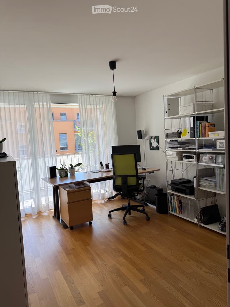 A modern, well-lit office space with a wooden floor, large window with sheer curtains, a desk with a computer setup, a comfortable office chair, and a metal shelving unit holding books, office supplies, and a printer.