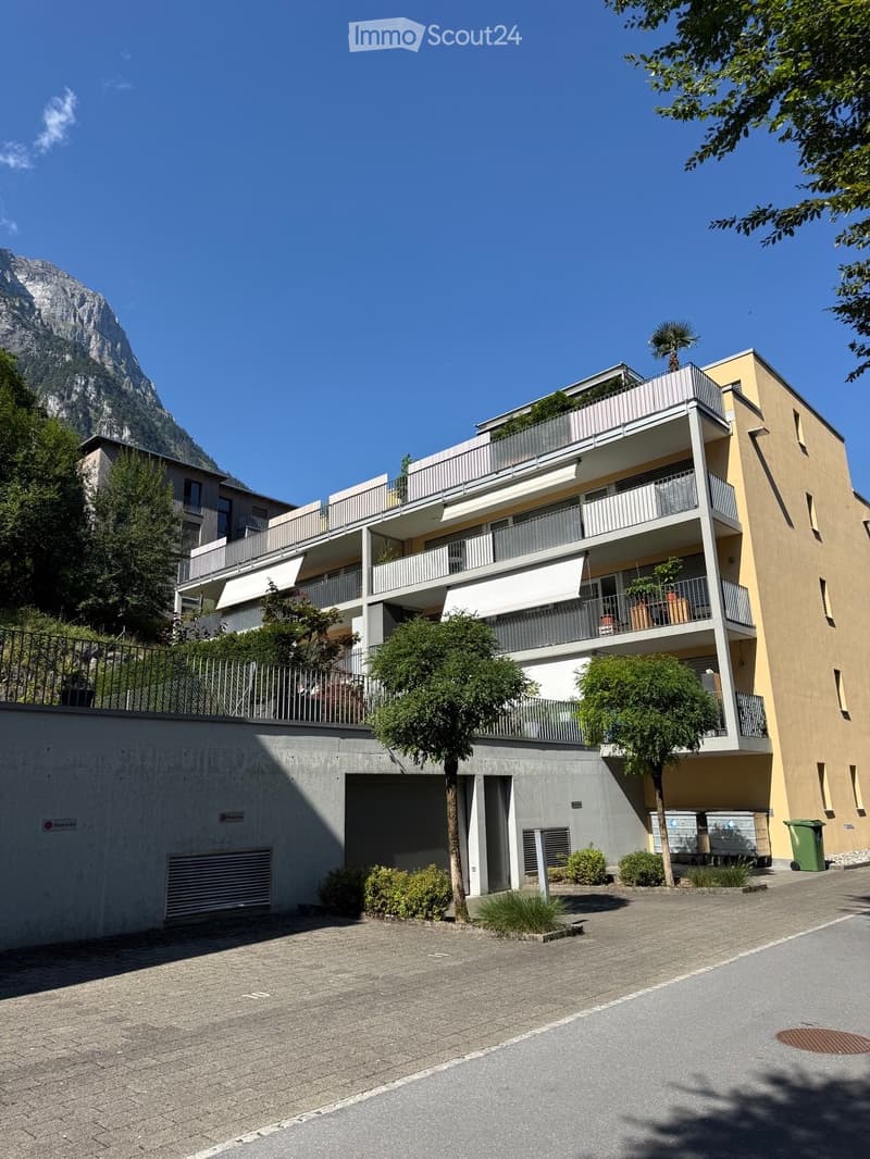 Modern multi-story residential building with balconies, surrounded by trees and a clear view of a mountain in the background.