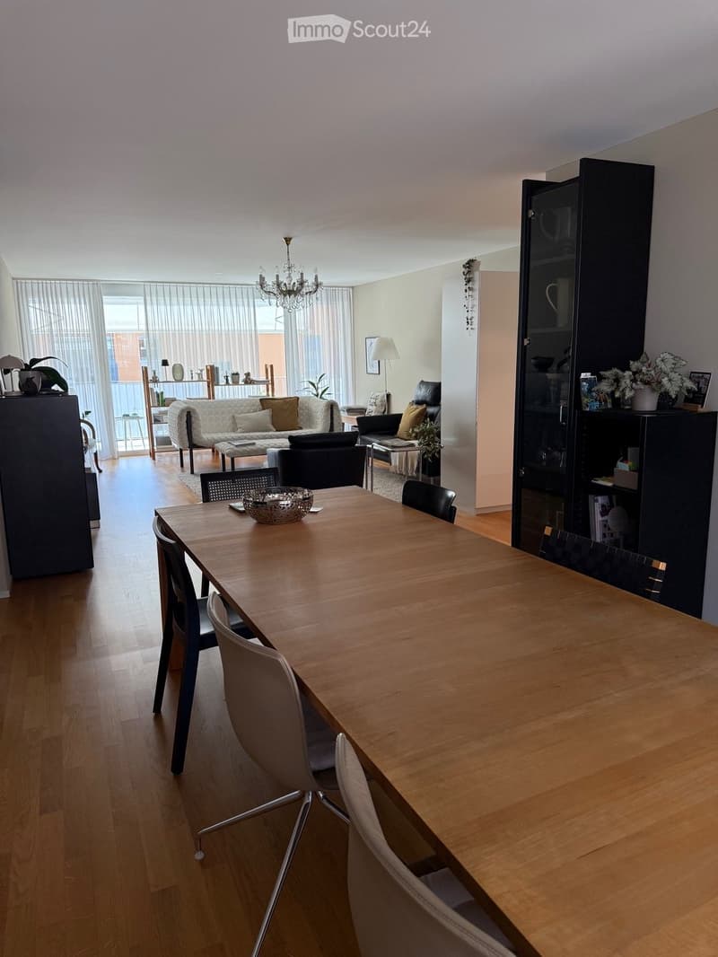 Modern dining and living area with wooden flooring, a long dining table with chairs, a sofa, armchairs, a bookshelf, and a chandelier.