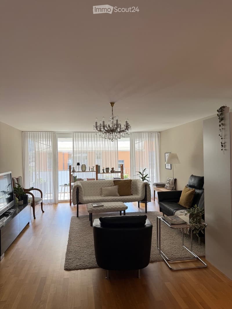 A modern living room with wooden flooring, a chandelier, a sofa, ottoman, chairs, a coffee table, a TV, and decorative plants.