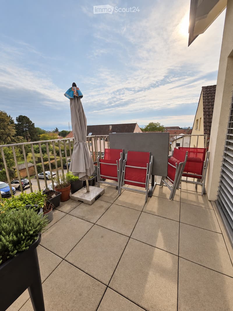 Balcony with tiled floor, red chairs, umbrella, and potted plants, overlooking a residential area with trees and parked cars.