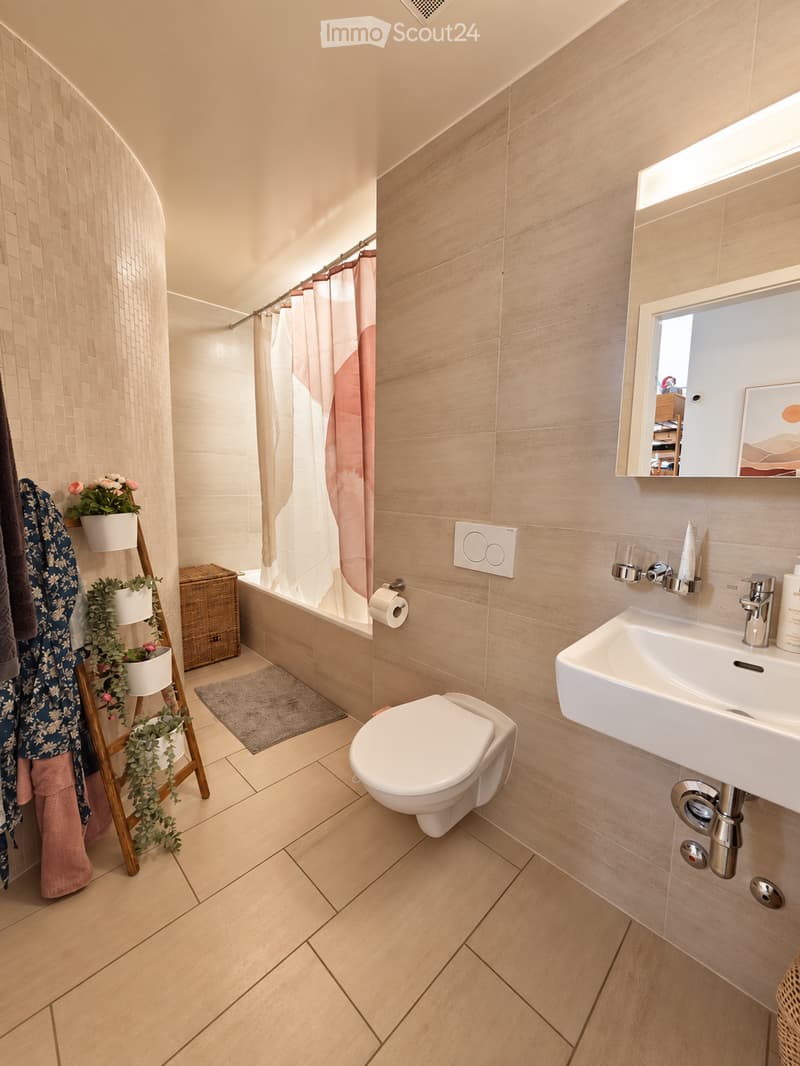Modern bathroom with light wood tiles, a white toilet, sink, bathtub, and a shower area with a pink curtain. Includes a mirror, towel rack, and decorative plants.