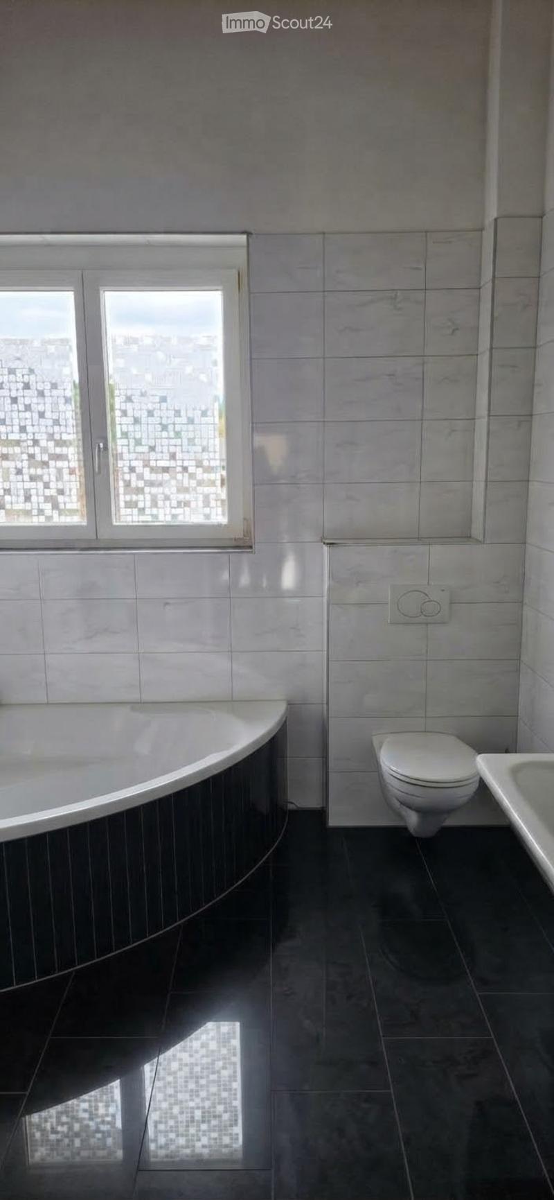 Modern bathroom with a bathtub, toilet, and sink. The walls and floor are tiled in a light grey marble pattern, and there is a window with frosted glass panes.
