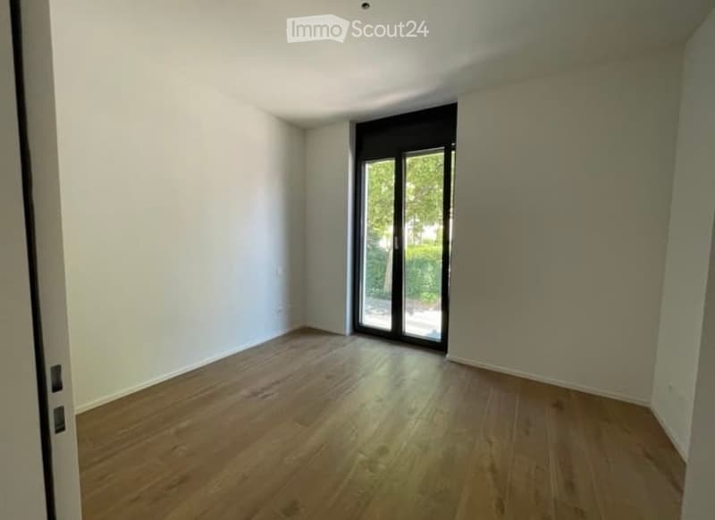 Empty room with light-colored walls and wooden flooring, featuring a large sliding glass door leading outside.