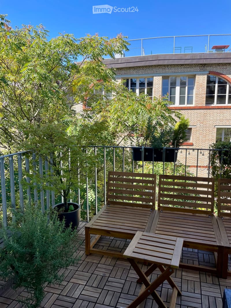 Wooden deck with two benches, a small table, and a potted plant, surrounded by greenery and a metal railing.