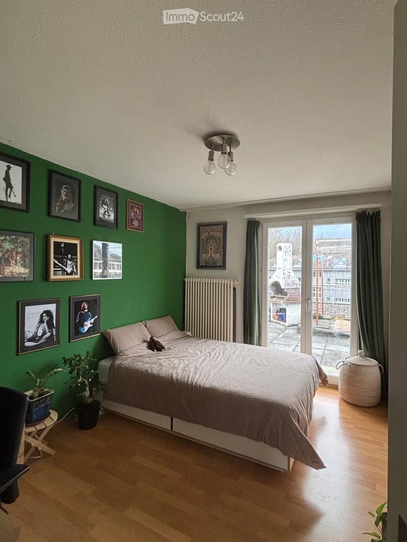 A modern bedroom with a large bed, wooden flooring, green accent wall, and several framed pictures. Features include a radiator, a small stool with a potted plant, and a large window with a view of the city.