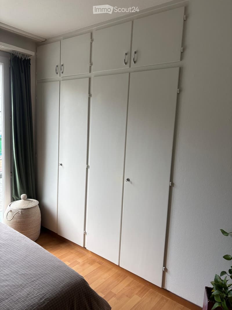 Bedroom with a large white wardrobe, a neatly made bed, a wicker basket, and a small plant in a pot.